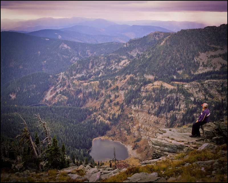 Wide perspective of Jim Cyr sitting on a ledge at the top of Eagle Peak looking out over Hub Lake and the vast Western Montana Wilderness of Mineral County.